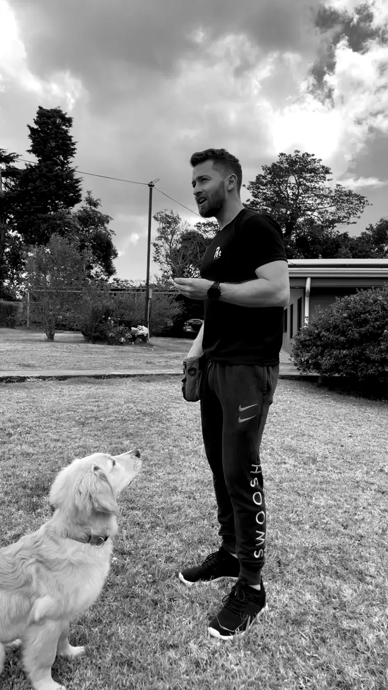 Jairo Sánchez de pie en un jardín, y un perro joven de pelo largo sentado a su lado, mirándolo atento, en blanco y negro.