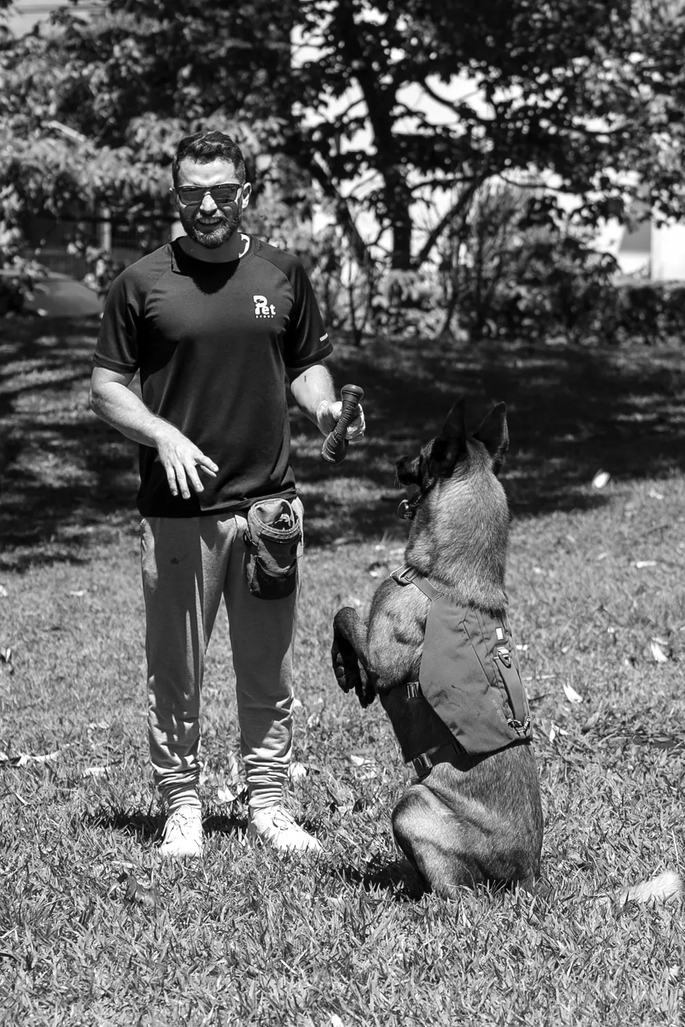 Jairo Sánchez sonriendo, con un juguete en la mano, frente a un pastor belga malinois sentado sobre las patas traseras que lo mira atento, en blanco y negro.