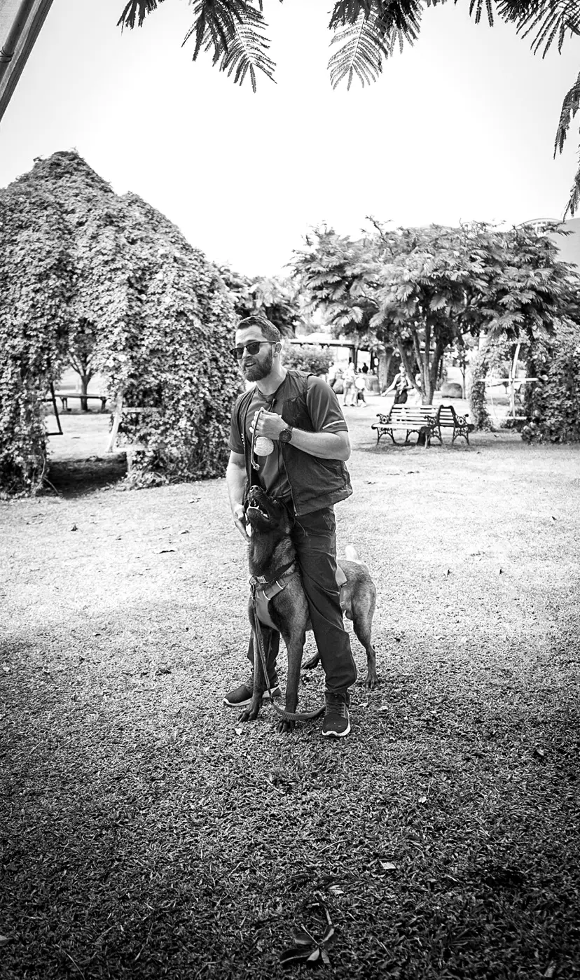 Jairo Sánchez de pie en un parque, con un pastor belga malinois tranquilo a su lado, en blanco y negro.