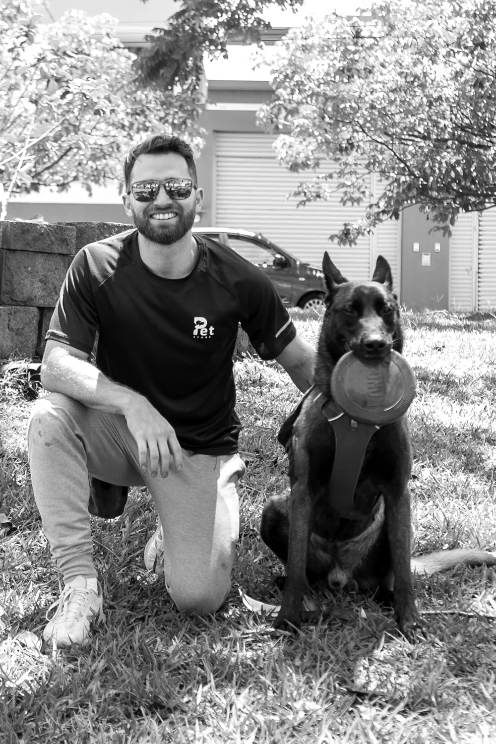 Jairo Sánchez agachado en el zacate, sonriendo, junto a un pastor belga malinois sentado con un disco en la boca, en blanco y negro.